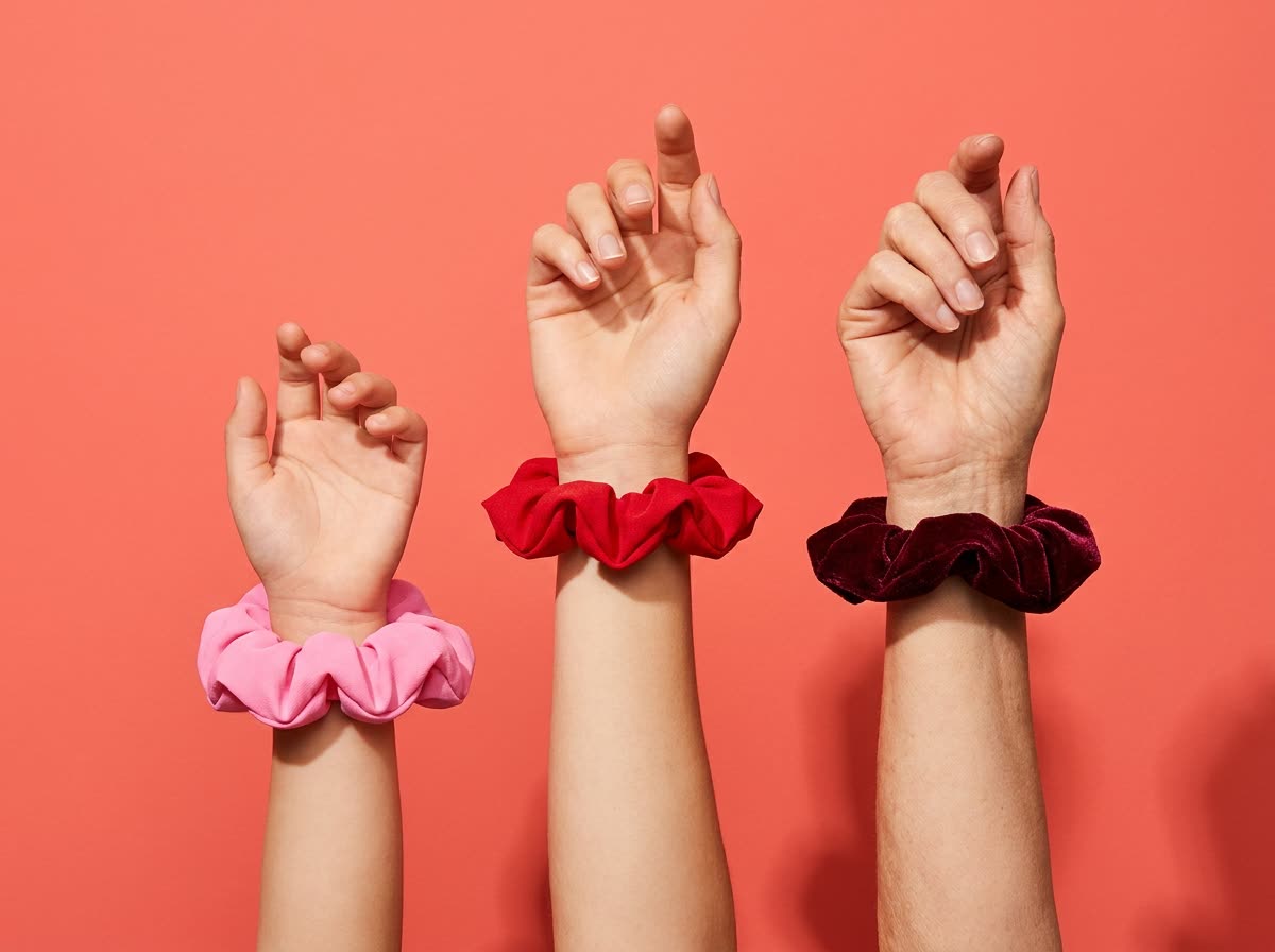 Three raised wrists — child, woman, older woman — each wearing a scrunchie
